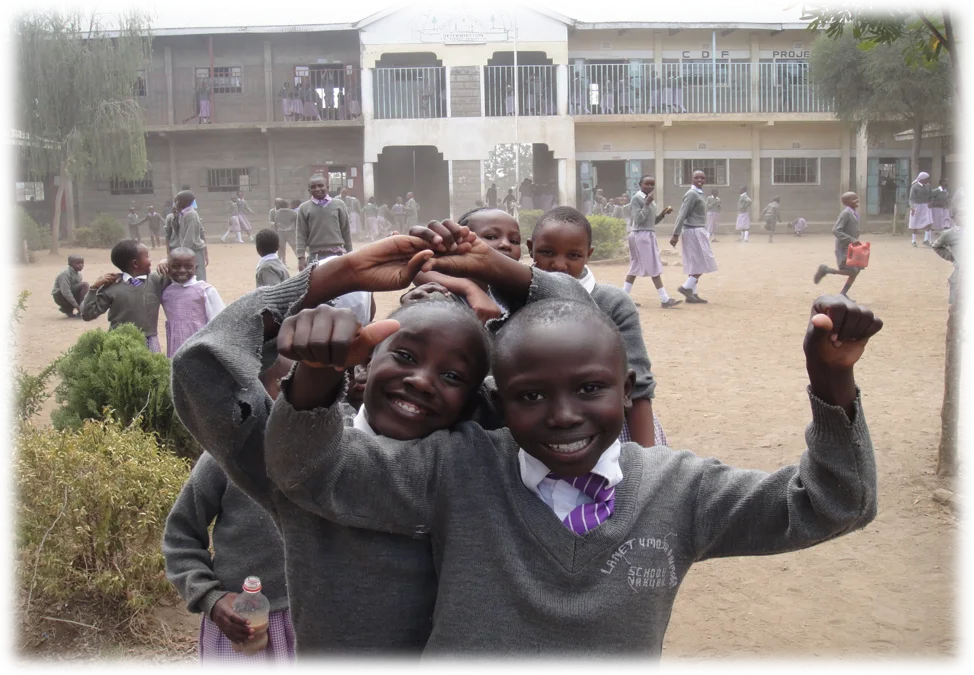 A group of smiling Kenyan schoolchildren in grey sweaters and purple-striped ties crowd playfully toward the camera, giving thumbs-up and making cheerful gestures. Behind them, more pupils in grey and lilac uniforms play in a sandy schoolyard in front of a two-story school building.